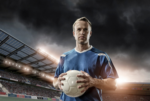 professional soccer player holding ball in full floodlit stadium under dramatic stormy sky at sunset
