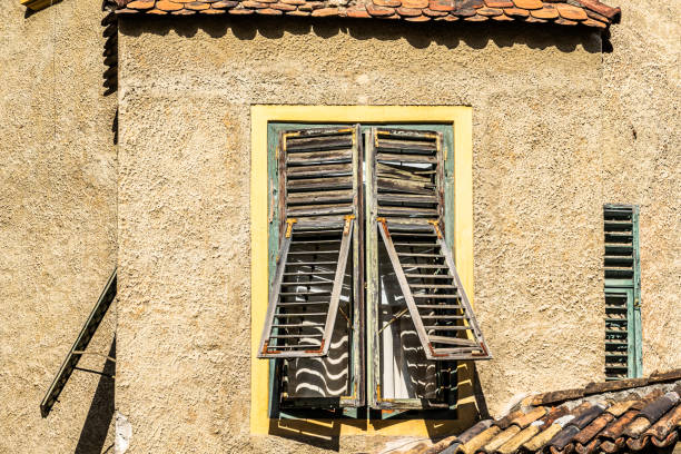 A window with a broken and dirty white shutter. The window trim is made of wood. The blind is broken and is falling to one side. It has a forged security grille. Villaviciosa de Odón-Madrid(Spain)
