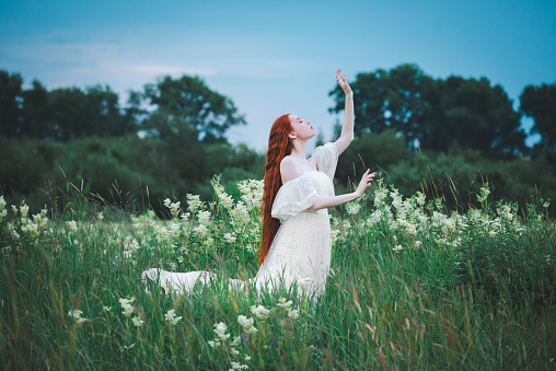 A red-haired sensual woman in a white dress of the 19th century on a background of a green field. The concept of lightness and freedom. The bride is outdoors in the summer.