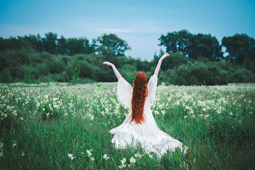 A red-haired sensual woman in a white dress of the 19th century on a background of a green field. The concept of lightness and freedom. The bride is outdoors in the summer.