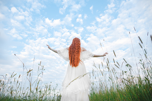 A red-haired sensual woman in a white dress of the 19th century on a background of a green field. The concept of lightness and freedom. The bride is outdoors in the summer.