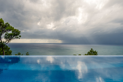 An ominous sky looms over a serene and calm ocean, featuring a subtle infinity pool in the foreground with a distant small tornado.