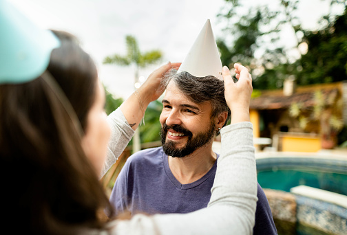 Woman helping a smiling male friend put on a party hat for a birthday party outside on a patio at home