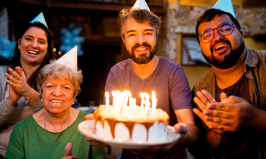 Portrait of a smiling group of family and friends singing and clapping while holding a candlelit birthday cake outside on a patio