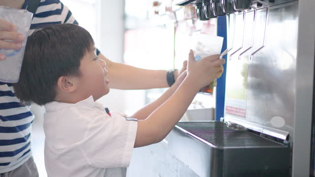 A little Asian boy presses a cold drink dispenser by himself in a Shabu restaurant.