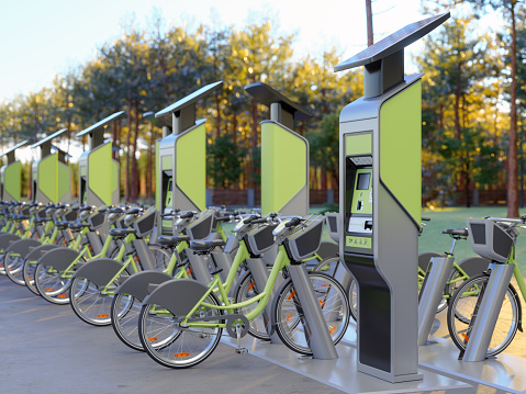 Row Of Bicycles In Parking Station With Blurred Parkland Background. Bicycle Sharing System