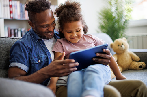 Couple using a digital tablet. They are sitting on a sofa with a modern kitchen behind them. They are attractive, smiling and happy.