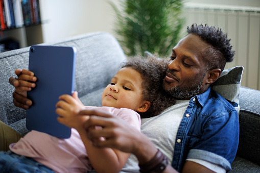 Happy African American father and adorable mixed race daughter using digital tablet in the living room at home