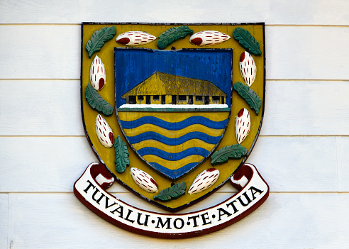 Vaiaku, Fongafale island, Funafuti Atoll, Tuvalu: Tuvalu coat of arms / seal, façade of the government headquarters - The coat of arms of Tuvalu is a shield with a golden border, which is decorated in a pattern with eight mitre shells and eight banana leaves. The shield itself shows a maneapa meeting house beneath a blue sky on green grounds. Beneath the ground are stylised depictions in blue and gold of ocean waves. The motto is 'Tuvalu mo te Atua', Tuvaluan for \