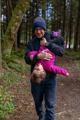 An active grandfather playfully lifts his giggling, two year old Eurasian granddaughter upside down as they hike through a forest in Oregon. The multi-generation family is on a camping trip on a cool, spring weekend in the Pacific Northwest region of the United States.