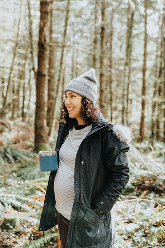 Active pregnant Eurasian woman wearing warm clothing, smiles and holds a hot beverage in a travel mug while hiking in the woods during a weekend away camping with her multi-generation family.