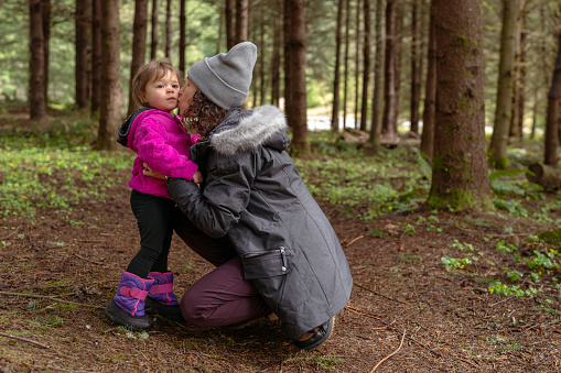 Eurasian pregnant mother of Asian and Hawaiian descent kneels next to her two year old daughter and gives the child a kissing on the cheek. The family is hiking through the forest while on a camping trip in the Pacific Northwest region of the United States.