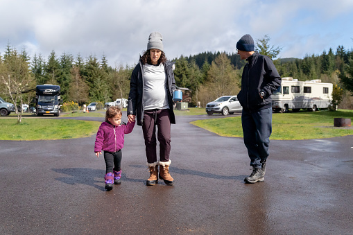 A pregnant Eurasian woman hold hands with her toddler daughter and takes a walk with her senior adult dad through a campground while enjoying a relaxing weekend away.