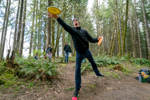 An active Eurasian man tees off while disc golfing along a forested course in Oregon while on a camping trip with his multi-generation family.