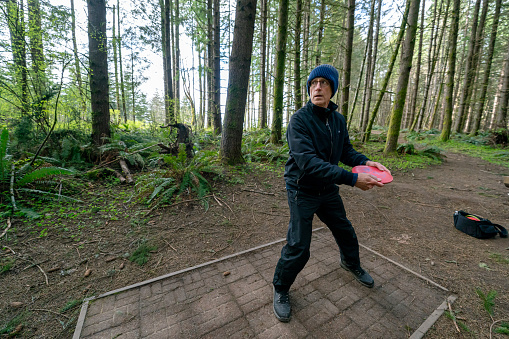 An active senior man tees off while playing a challenging disc golf course located in a forest in the Pacific Northwest region of the United States.