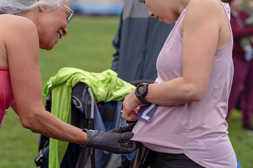 Cropped profile view of a pregnant Eurasian woman attaching a race bib to her shirt with her mother's help before running in a race.