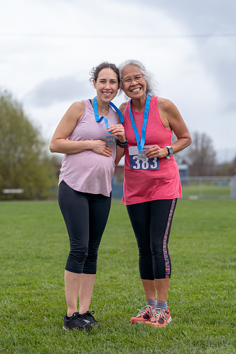 An active Eurasian woman who is pregnant rests her hand on her growing belly while posing with her fit senior adult mom after completing a half marathon race together. The mother and daughter are standing together in a grassy field and are holding their race medals.