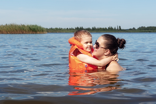 Portrait of happy little boy in orange swimming vest hugging his mother in lake water.