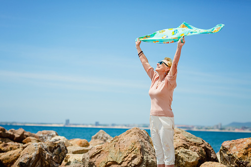 Happy mature woman smiling and having fun at beach. Summer portrait of senior woman walking on beach with a blue scarf