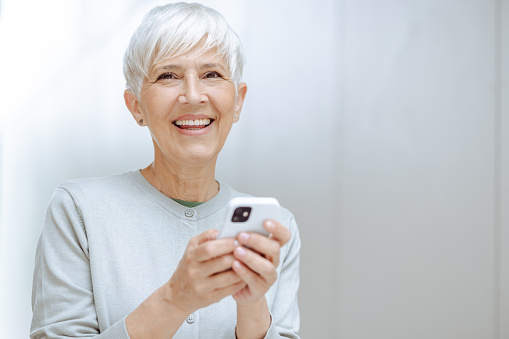 Shot of a happy senior woman with short gray hair in front of the gray background. She is holding smart phone and looking at the camera