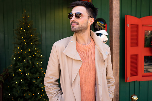 Handsome man wearing sunglasses and trench coat standing outside near Santa house on Christmas market looking away and smiling. Portrait young bearded male person