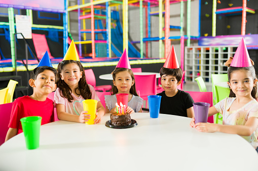 Cheerful group of children celebrating their friends birthday party with a cake wearing hats in a beautiful indoor playground