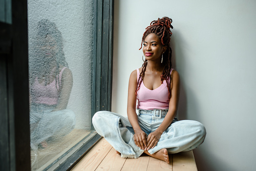 Attractive smiling African woman wearing ribbed tank top and jeans sitting cross-legged on the windowsill and looking through the window. Urban life concept, daydreaming. Planning future concept