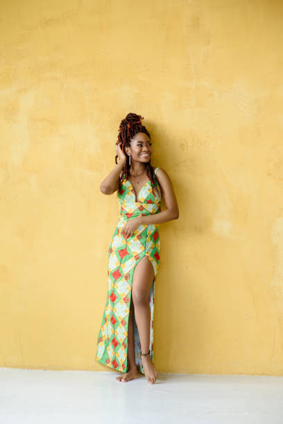 istock The attractive African woman joyfully smiling while looking away, wearing a long multicolor ornament high slit dress, standing barefoot in front of a yellow wall background with copy space. 1570532371