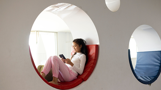 A cute   girl in a light spring dress sitting on a round window and using a smartphone  with bright sunlight outside , resting inside a circular window