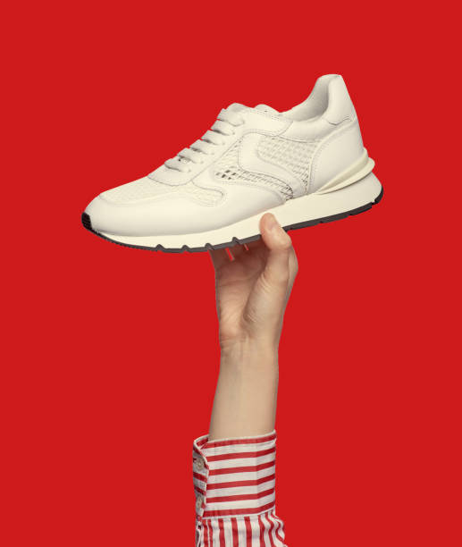 istock A woman's hand holding a modern white sneaker, isolated on a red background. Sport and active lifestyle.  Outlet marketing poster. Summer shoe sale campaign 1570541303