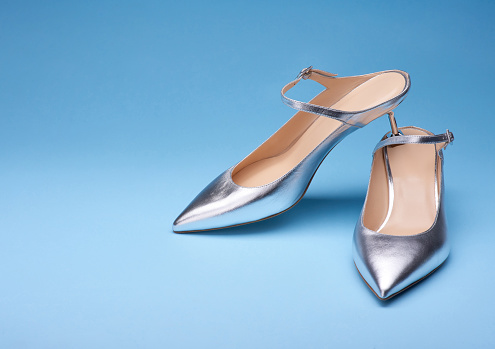 Bold silver leather pumps with open backs and clasps on the straps standing one on another isolated on blue background. Copy space for text.  Mock-up for design advertising for a shoe store