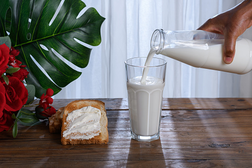 Glass of milk, bottle pouring milk into a glass, toast on a rustic wooden table in front of a window with a white curtain.