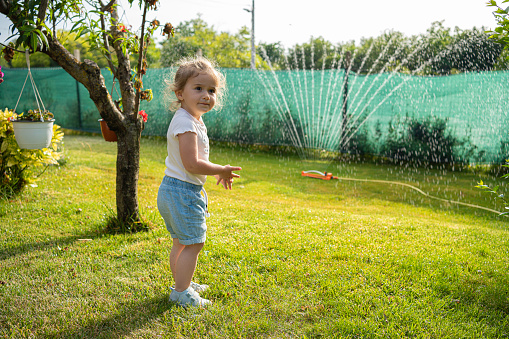 Cute little girl playing with water from garden hose sprinkler in a backyard.