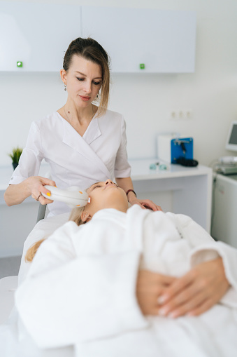 Vertical portrait of female beautician performing photo rejuvenation cosmetology procedure for woman in beauty clinic. Concept of non-surgery cosmetology, professional skincare, beauty treatment.