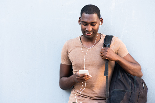 Portrait young African American man with bag looking at mobile phone while listening to music