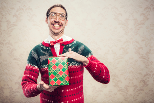 A man in a knit reindeer Christmas cardigan button up sweater, complete with matching red bow tie and a classy mustache shows off a wrapped gift he's ready to give as a present.  Damask style vintage wall paper in the background.  Horizontal with copy space.