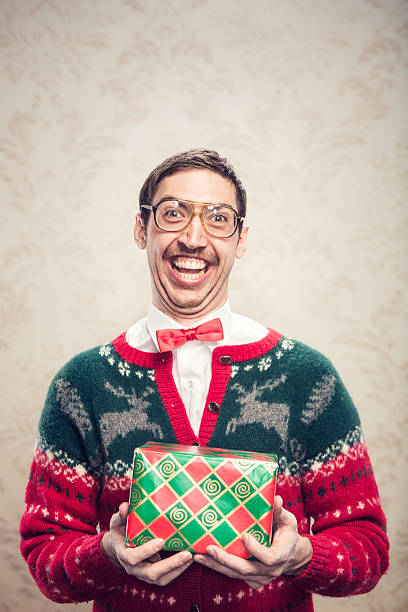 Christmas Sweater Nerd A man in a knit reindeer Christmas cardigan button up sweater, complete with matching red bow tie and a classy mustache shows off a wrapped gift he's ready to give as a present.  Damask style vintage wall paper in the background.  Vertical with copy space. christmas nerd sweater cardigan stock pictures, royalty-free photos & images