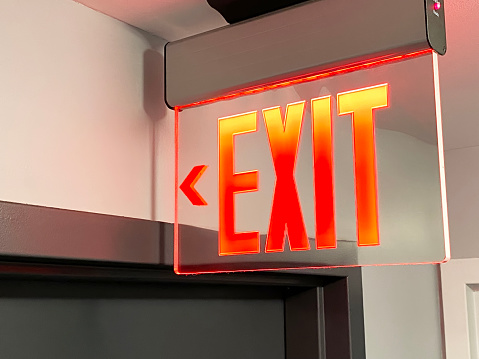 Focus at green electric emergency exit sign on ceiling with blurred background of many people inside of shopping mall.