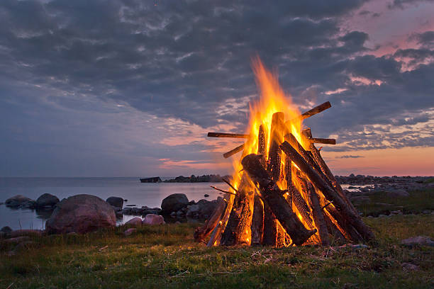Blazing bonfire near a body of water at dusk Bonfire on the coast in a white Nordic summer night.  bonfire stock pictures, royalty-free photos & images