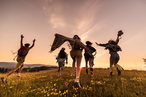 Rear view of playful backpackers having fun while running on a meadow at sunset. Copy space.