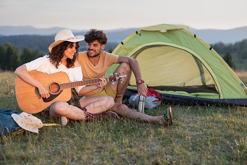 Happy woman playing a guitar for her boyfriend during camping in nature. Copy space.