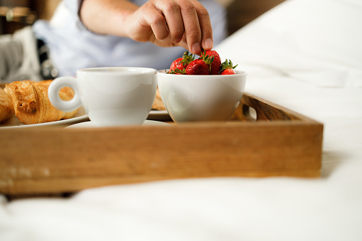 Close up of unrecognizable man taking strawberry from a tray during breakfast in bedroom.