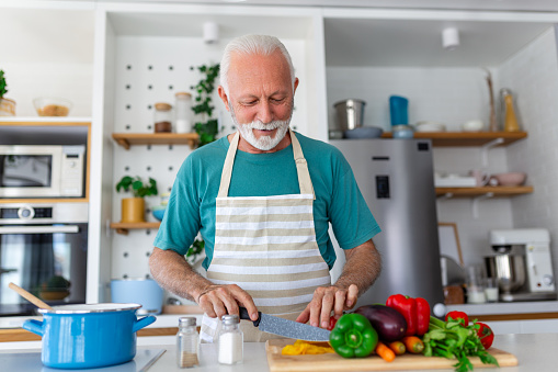 Happy retired senior man cooking in kitchen. Retirement, hobby people concept. Portrait of smiling senior man cutting vegetables