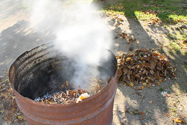 Rusty metal barrel with white smoke to burn the fallen leaves in the autumn season, with in the background a pile ready to burn. Focus on the barrel without people.