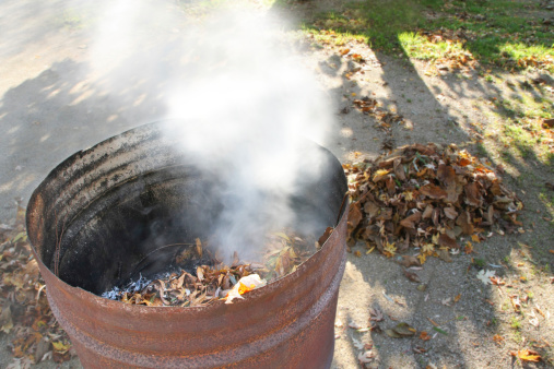 Rusty metal barrel with white smoke to burn the fallen leaves in the autumn season, with in the background a pile ready to burn. Focus on the barrel without people.