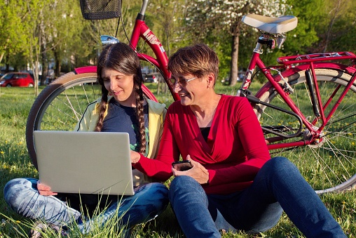 Mature and young woman sitting in park using laptop - mother and daughter with red bicycle and laptop