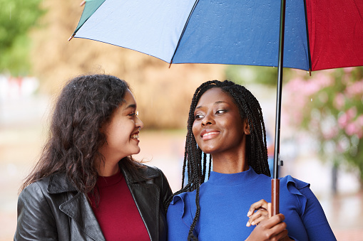 Multi-ethnic female friends smiling while walking outdoors with a colorful umbrella in a rainy day. Friendship concept.