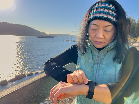 Asian senior woman checking health statistics on smart watch on pedestrian seawall, with ocean in the background.  North Vancouver, British Columbia, Canada.
