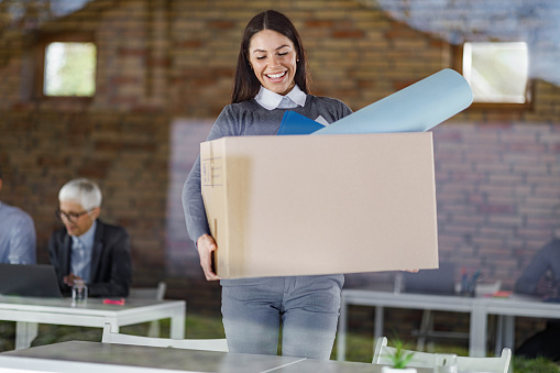 Happy female entrepreneur carrying her belongings after quitting from a job. The view is through glass.