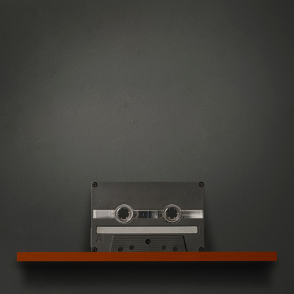Blank audio cassette on a wooden shelf against dark concrete wall with copy space.